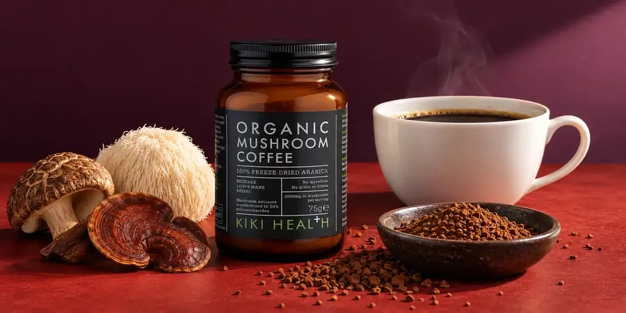 Sklenice KIKI Health Organic Mushroom Coffee vedle šálku kávy, misky s instantním práškem a plodnic hub na bordó pozadí